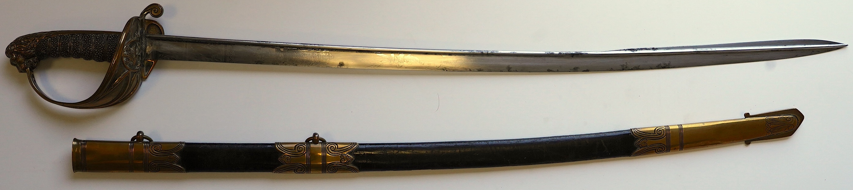 An early 19th century Royal Navy officer's sword with engraved blade, folding knuckle guard, lion’s head pommel and shagreen grip, leather scabbard with brass fittings, blade 73.5cm. Condition - fair, some wear overall.